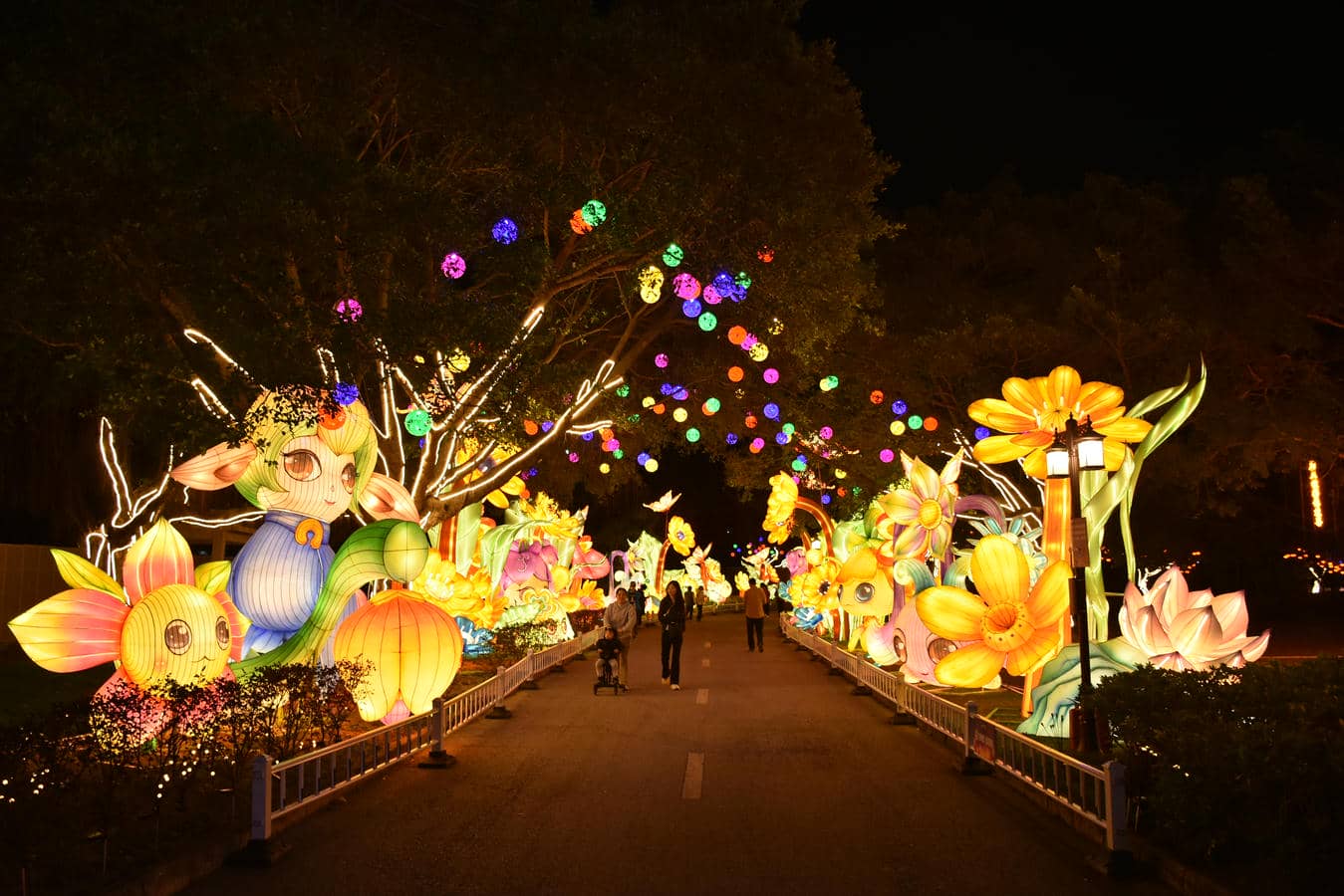 Visitors walk along an outdoor park light show route surrounded by colorful flower and animal lantern displays