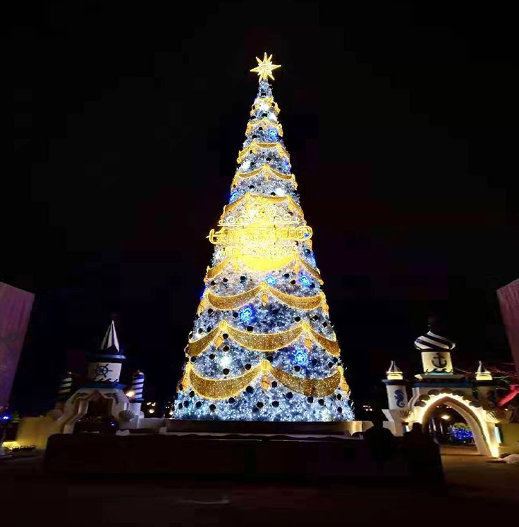 Tall outdoor Christmas tree adorned with blue and gold lights and ornaments, glowing against a dark night sky in a public display.

