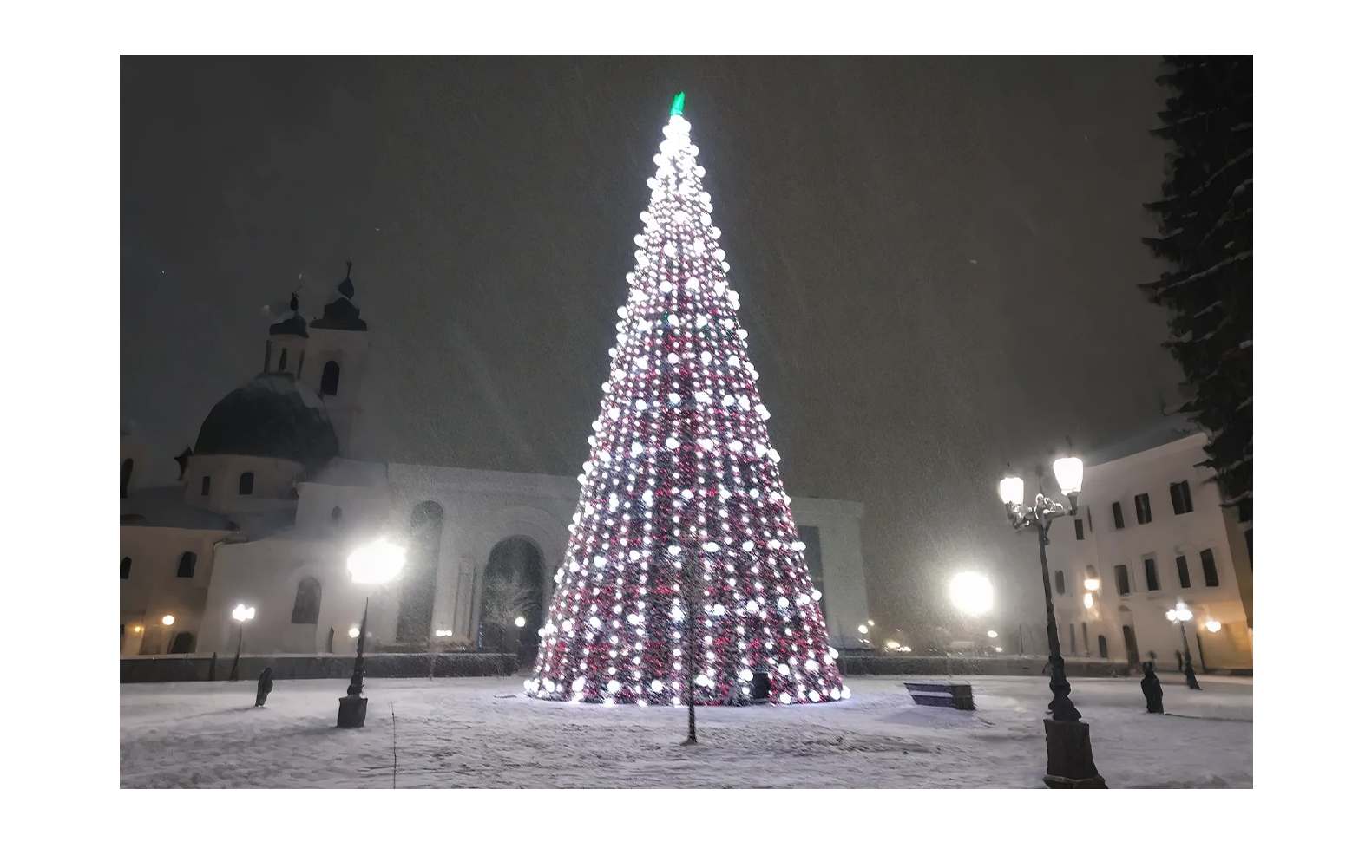 A majestic Christmas tree adorned with white and red lights stands prominently in a snowy square at night. The tree is surrounded by historic architecture, including a building with domed towers and arched windows, illuminated by street lamps. Snow gently falls, creating a festive and serene atmosphere.